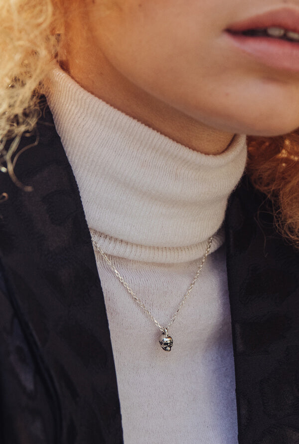 A women with blond hair wearing a white polo neck jumper and a black jacket. She is also wearing a silver small skull necklace.